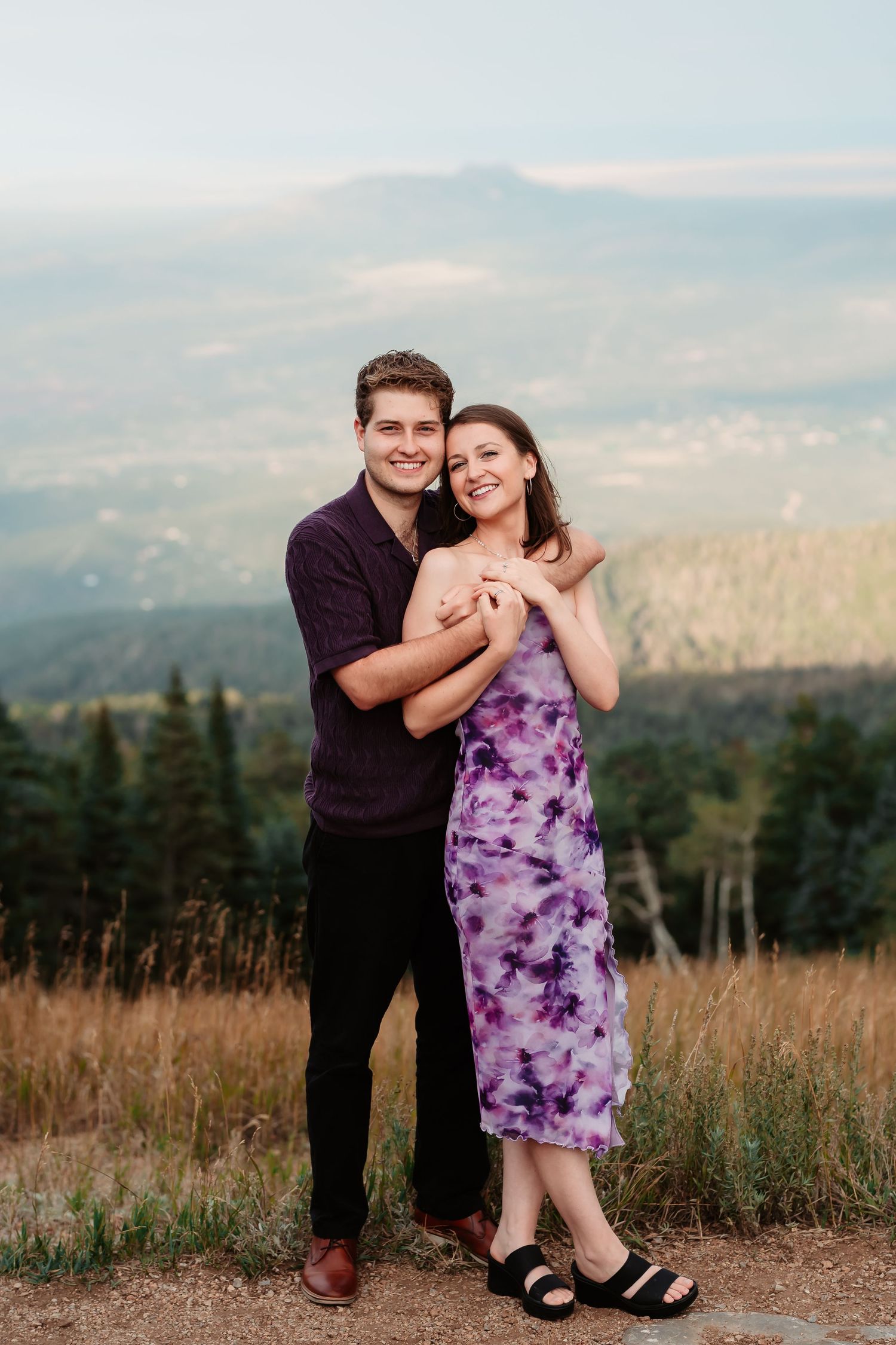 Romantic engagement photos in mountain setting with couple in purple dress and dark shirt.