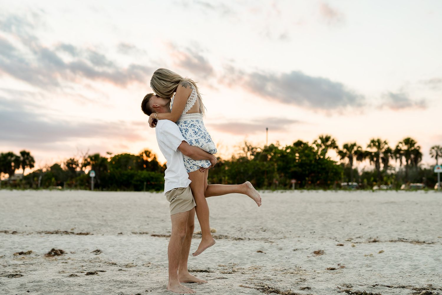 Couple sharing a romantic kiss on a sandy beach at sunset with palm trees in the background.