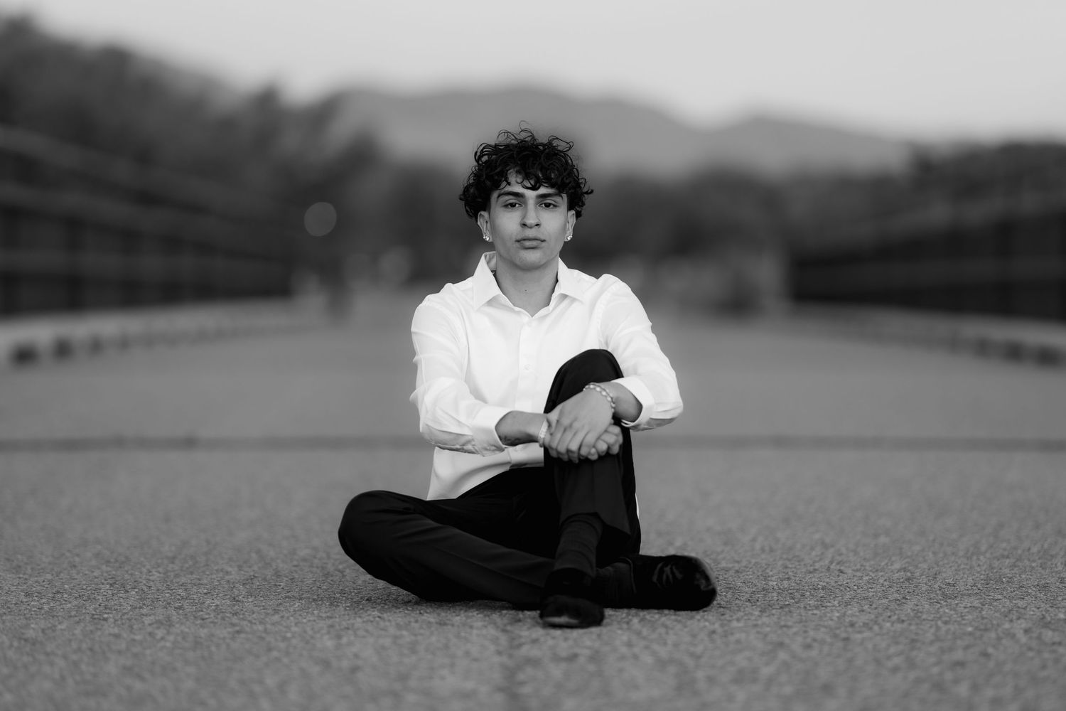 Black and white portrait of someone sitting cross-legged on a road wearing a white shirt and dark pants.