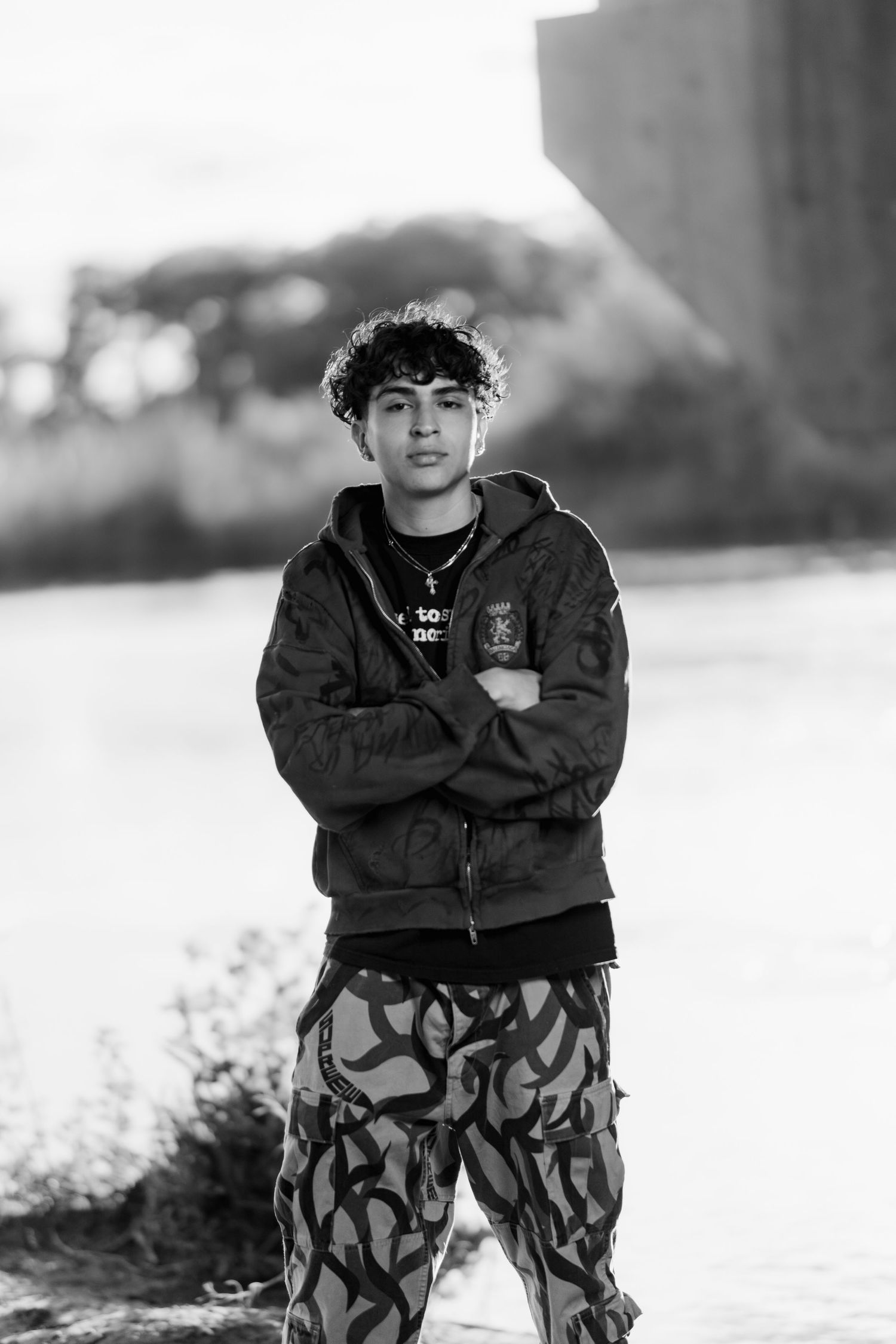 Black and white portrait of someone in a hoodie and patterned pants standing outdoors.
