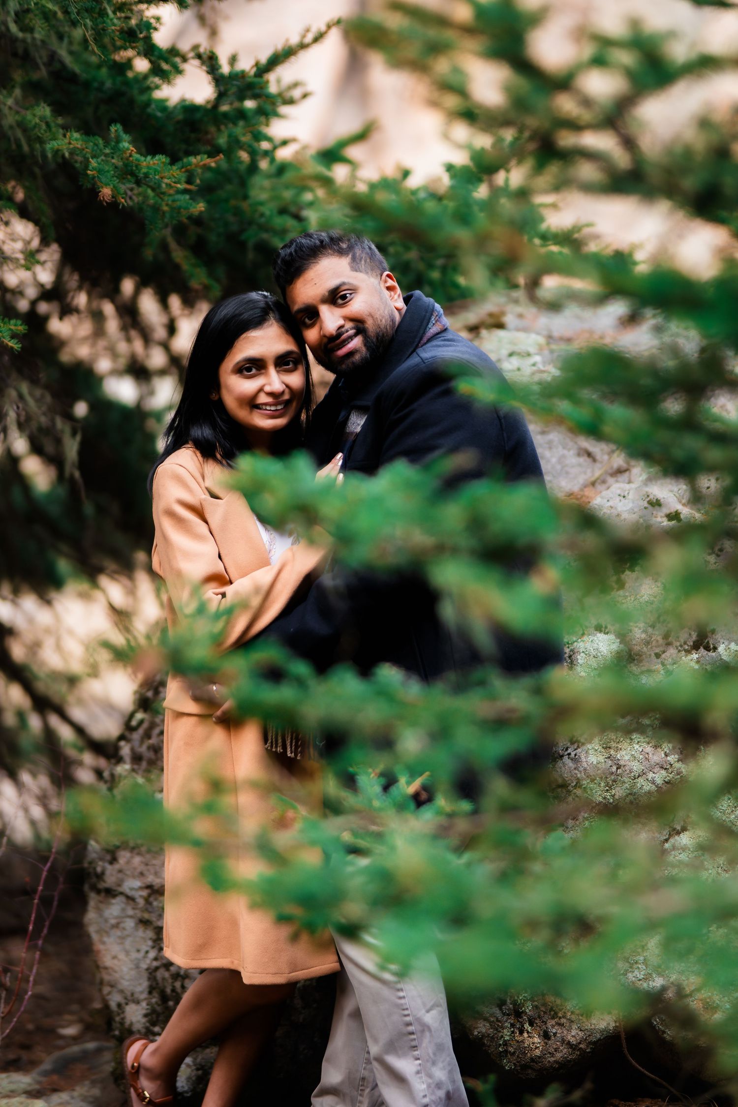 A couple shares a romantic moment between evergreen trees in a scenic outdoor setting.