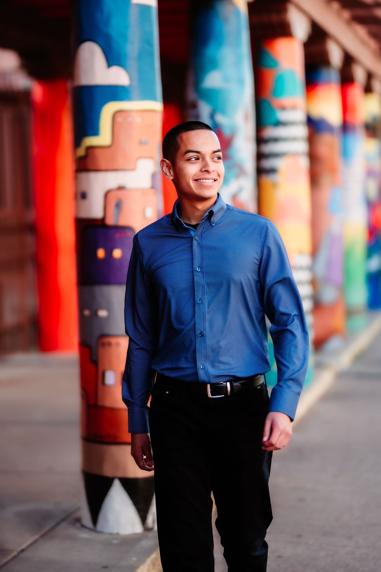 A person in a blue button-down shirt smiles while standing in front of colorful painted columns with geometric designs.
