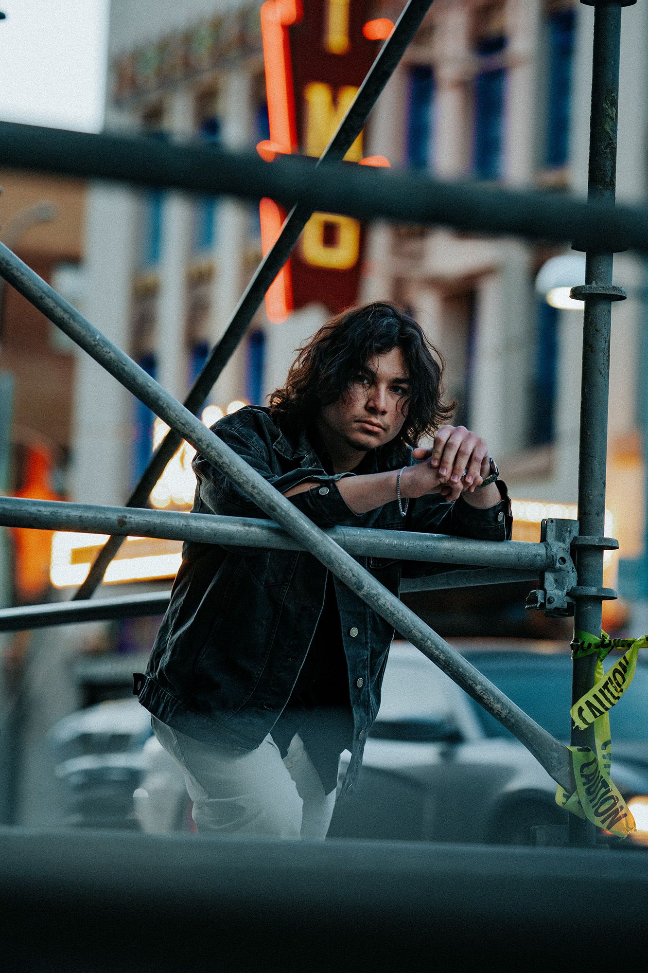 Person in dark clothing poses dramatically against metal railings in an urban setting at dusk.