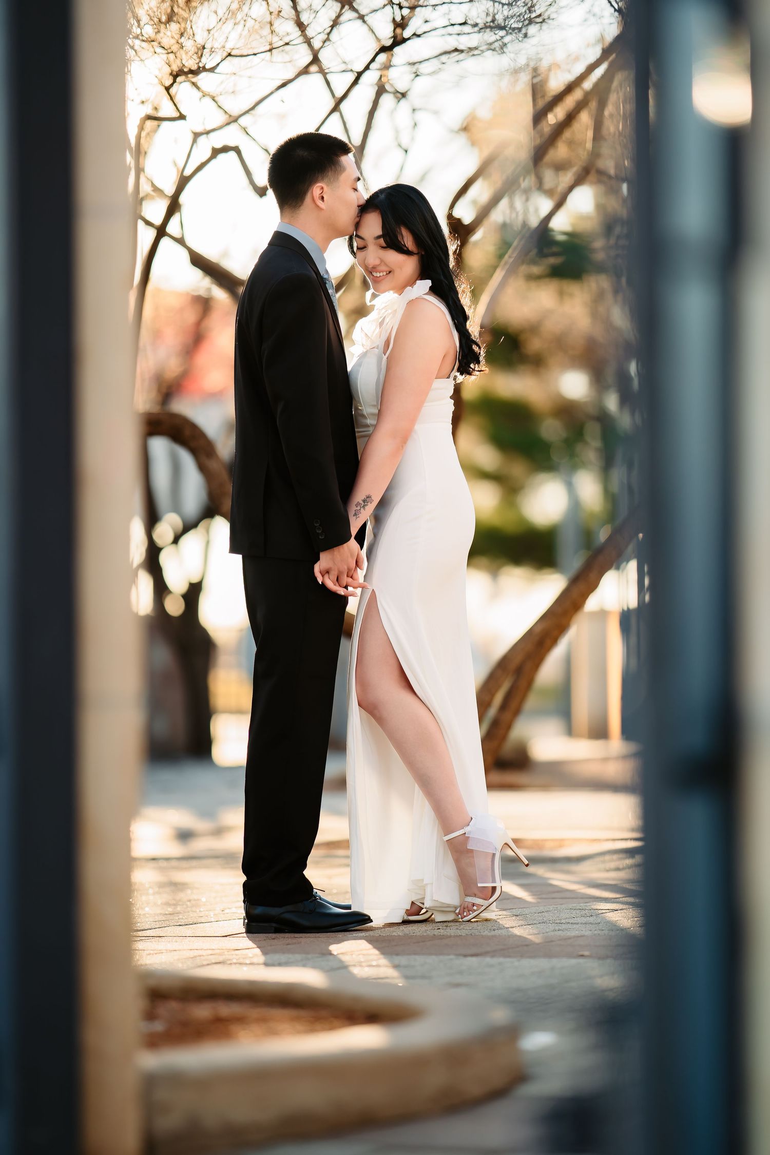 Bride and groom share a romantic kiss at sunset through a decorative iron gate in an outdoor garden setting.