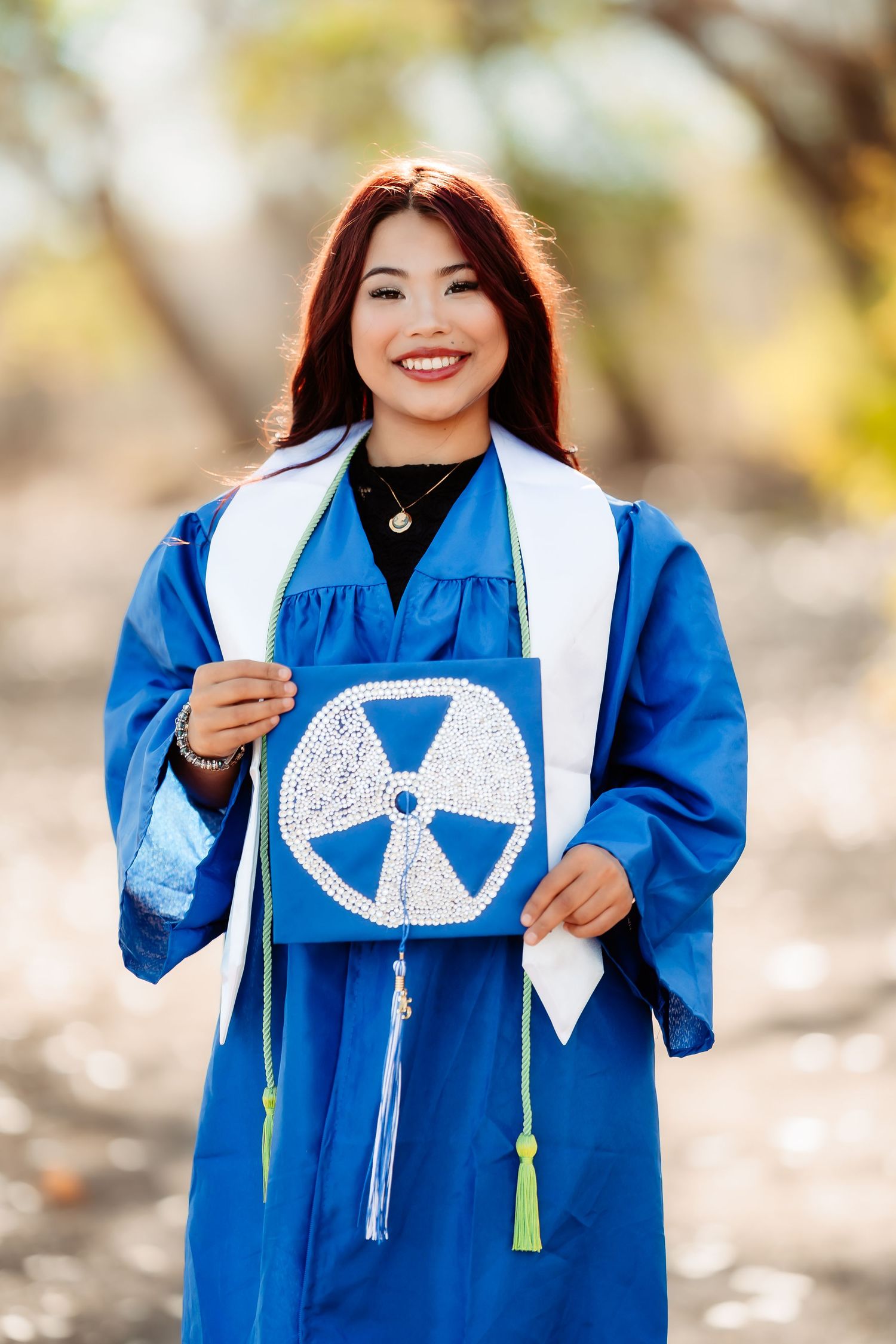 Graduate wearing a blue graduation gown and white stole with decorative designs poses for photos outdoors.