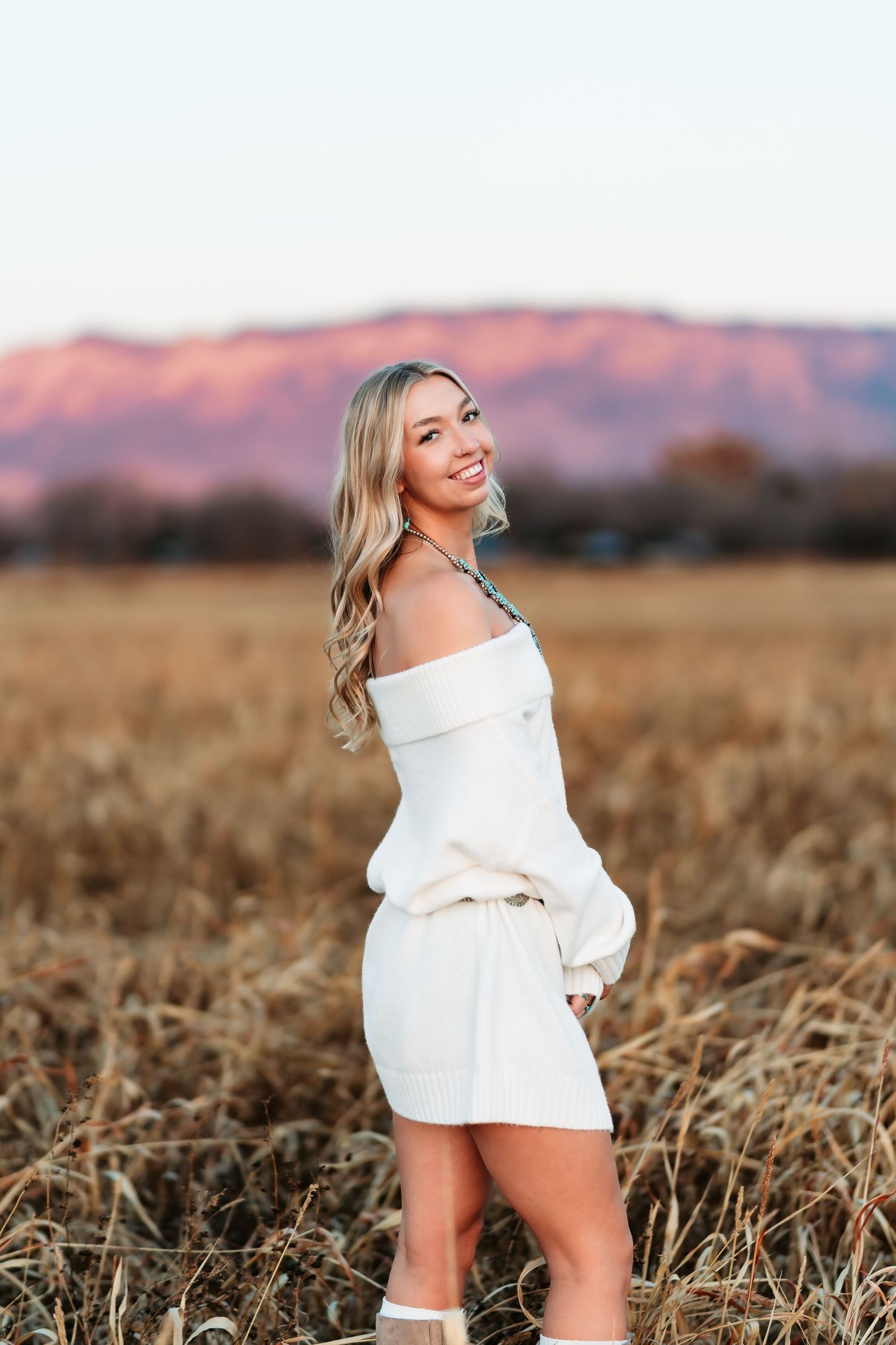 A person in a white outfit poses in a golden field at sunset with mountains visible in the background.