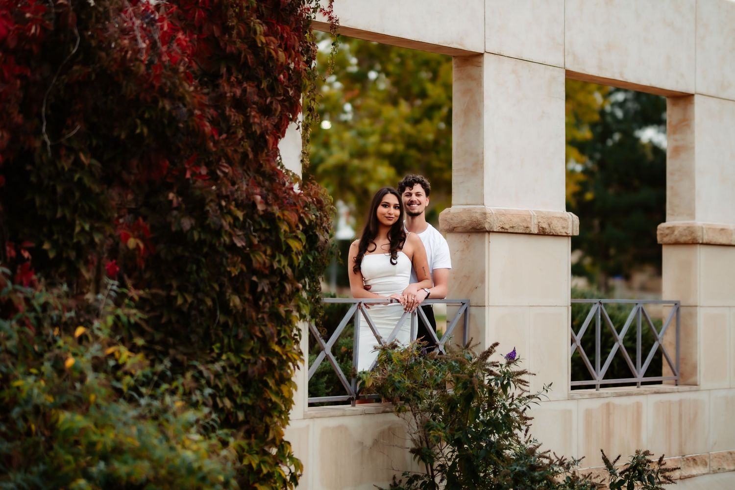 A couple in wedding attire stands together on a white stone balcony surrounded by autumn foliage.