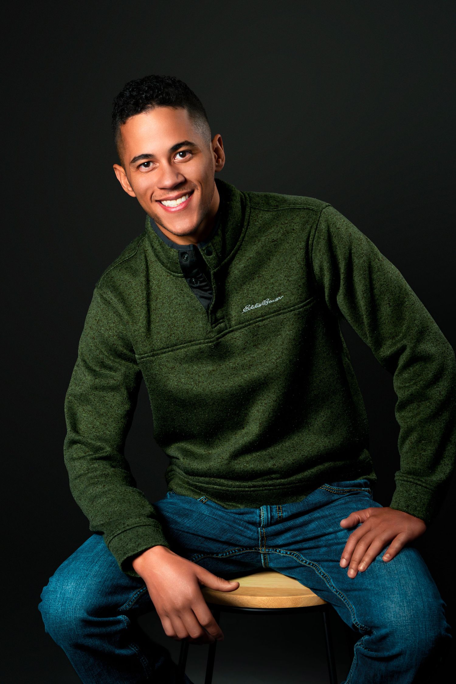Professional studio portrait of person in green fleece pullover and jeans sitting casually against dark background.