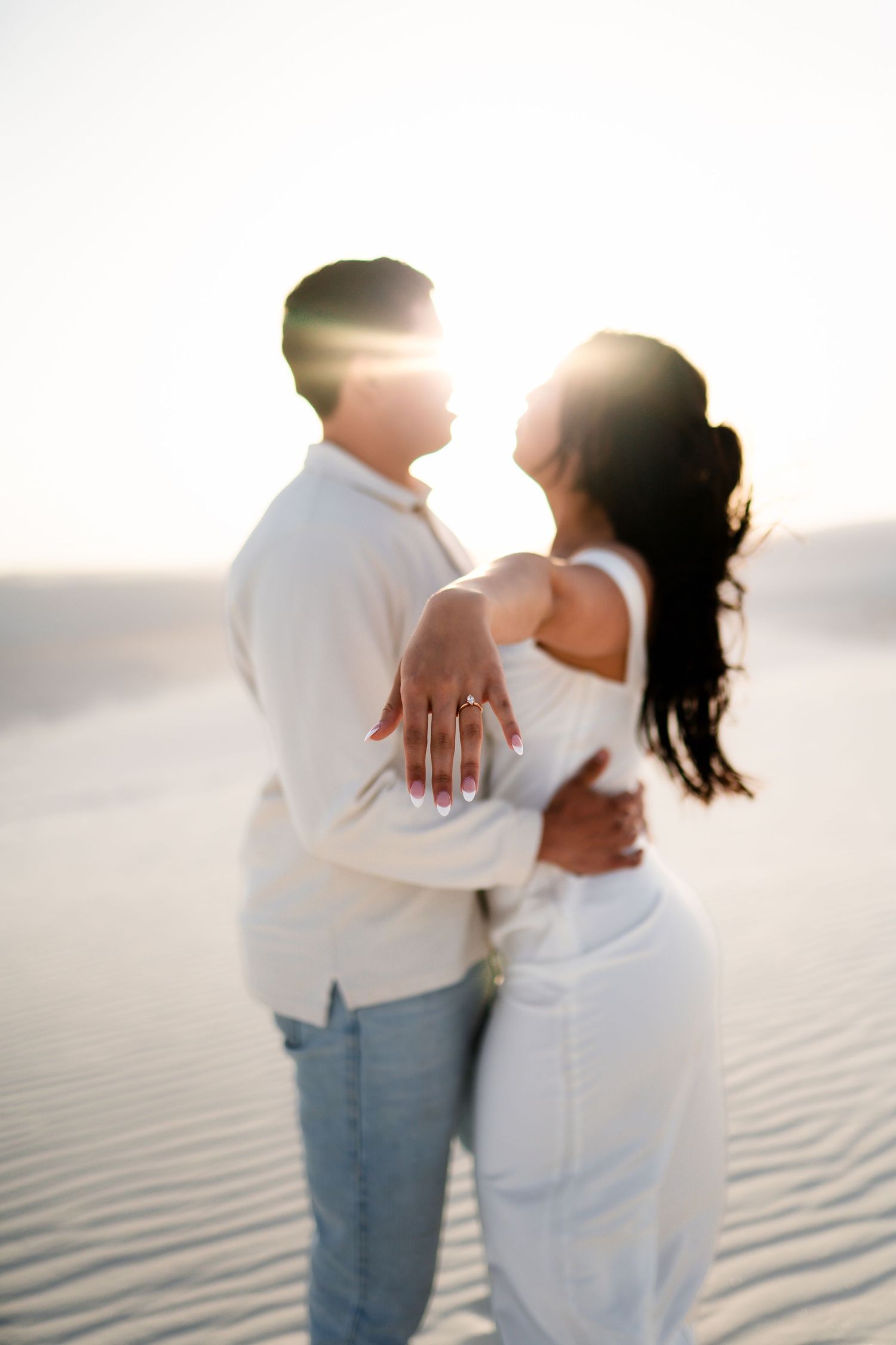 A romantic couple embraces on a sandy beach at sunset, with the woman showing off her engagement ring.