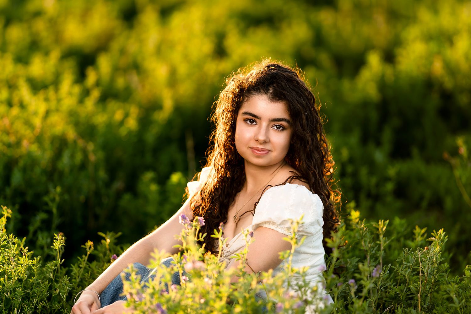 Young woman with curly hair sits among wildflowers in golden sunlight, wearing a white dress in a lush green field.