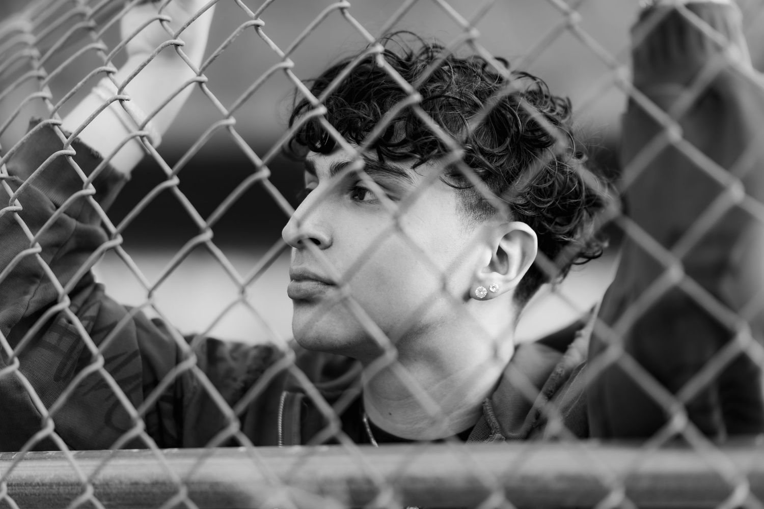 Artistic black and white shot through a chain-link fence showing a thoughtful pose.