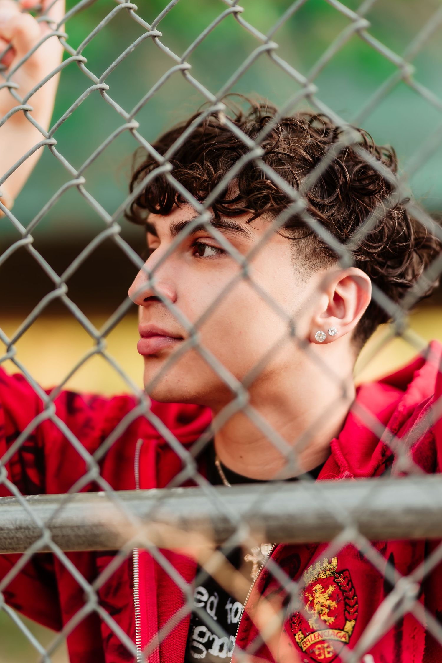 Through chain link fence shot of person in red hoodie outdoors with curly hair and an earring in profile view.