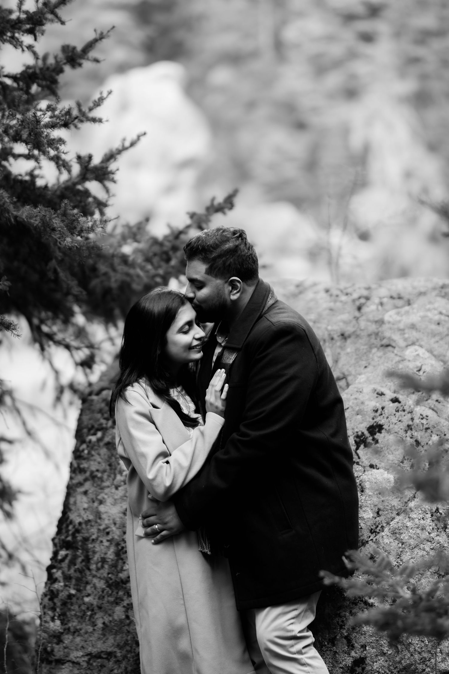 A romantic black and white photo of a couple embracing outdoors near an evergreen tree in a natural setting.