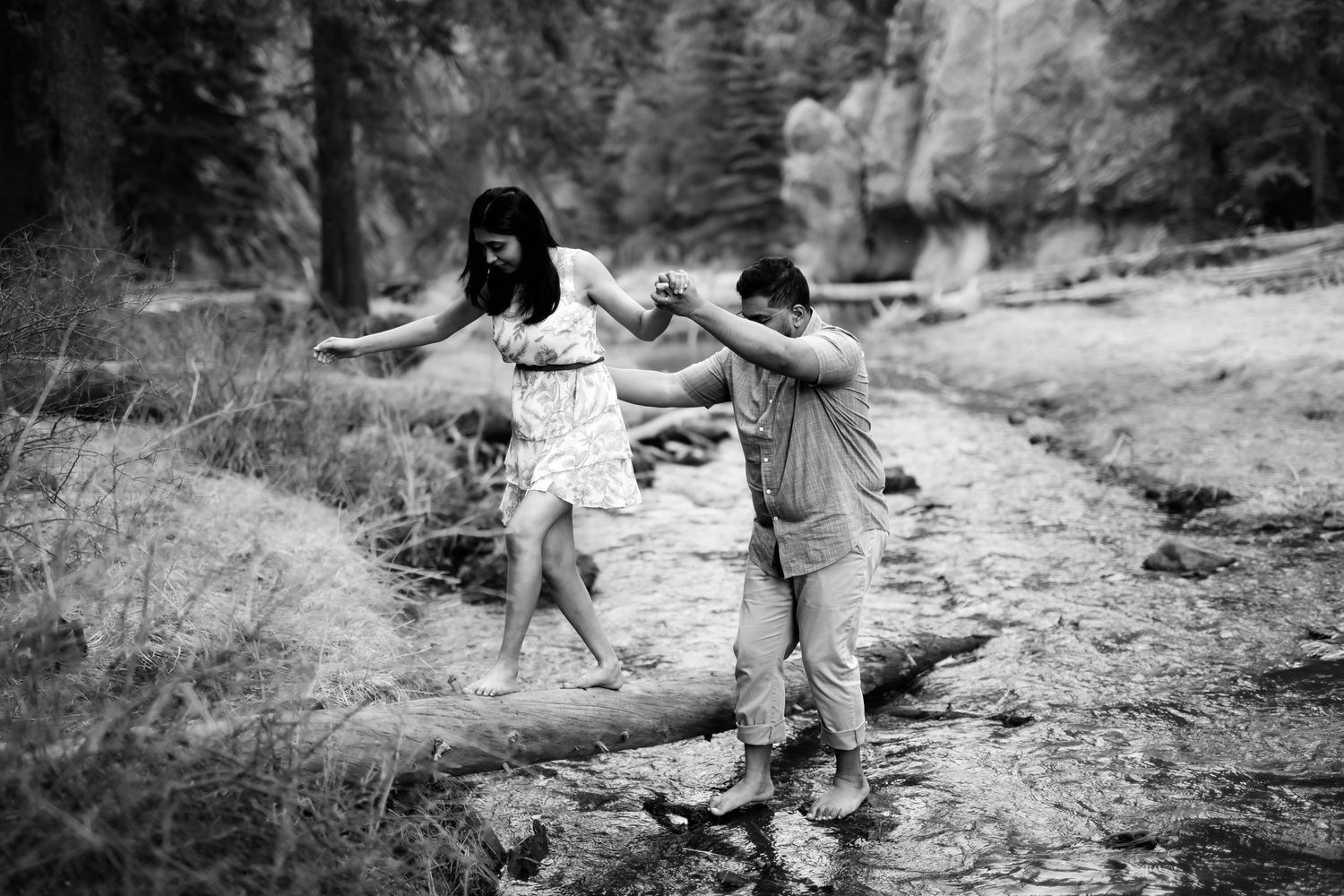 Two people carefully balance while walking on rocks near a river in a black and white nature setting.