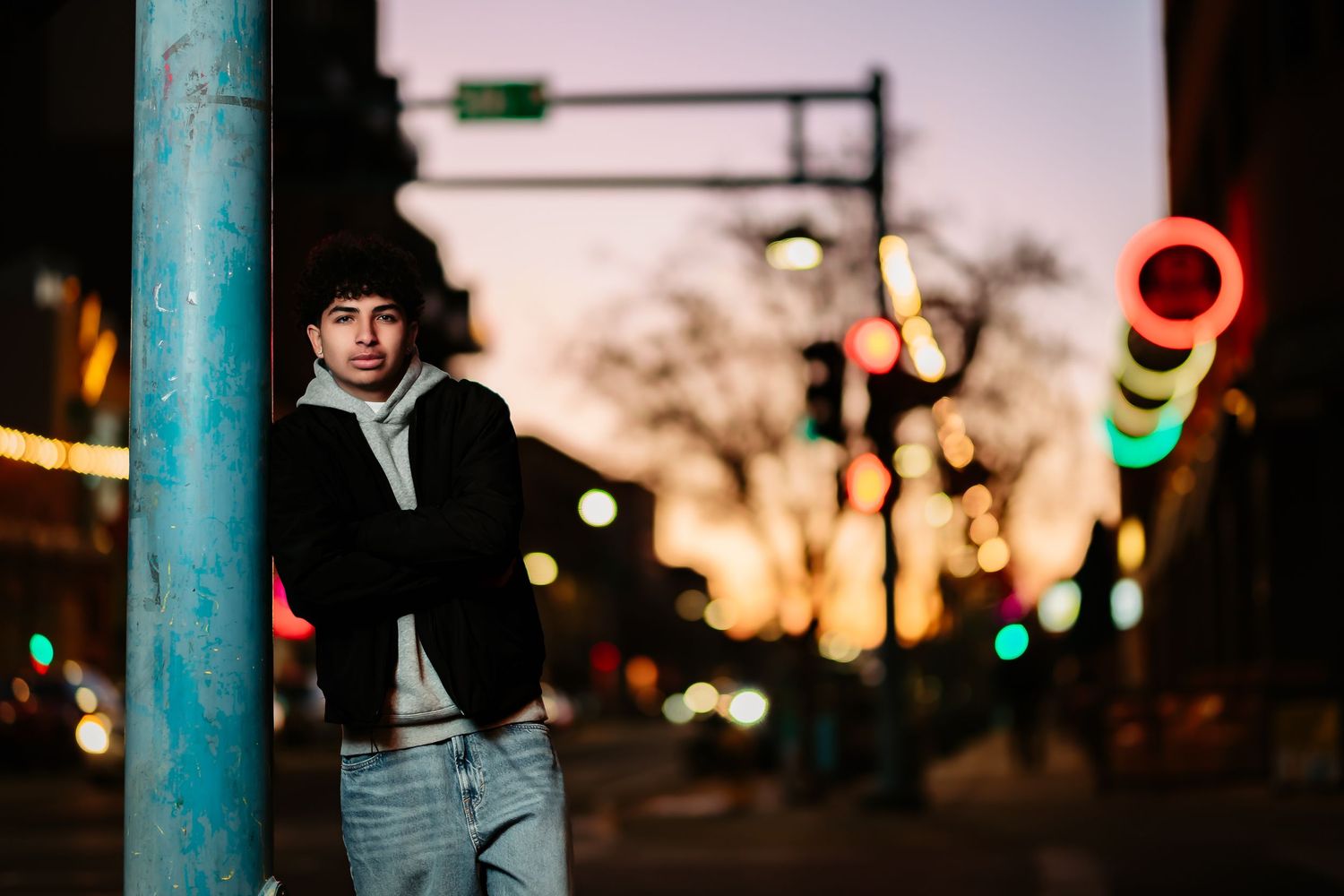 A person in a black jacket stands between blue poles at dusk with blurred city lights in the background.