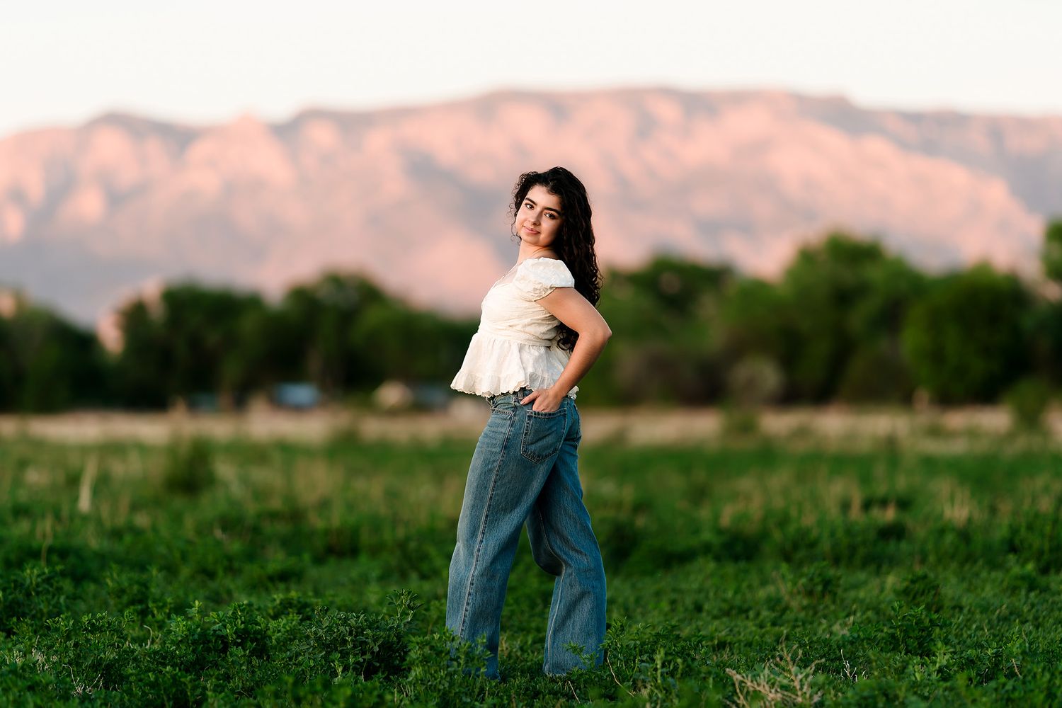 Young woman in white top and wide-leg jeans poses in green field with pink mountain backdrop at sunset.