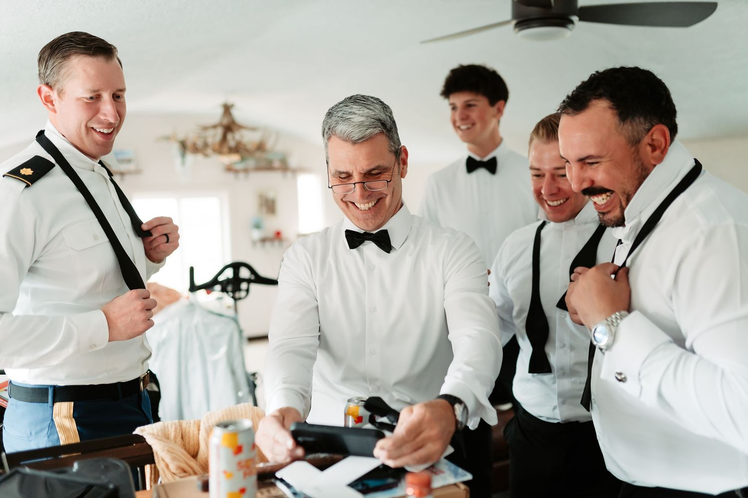 Groomsmen in white dress shirts and black bowties laugh together while getting ready for a wedding celebration.