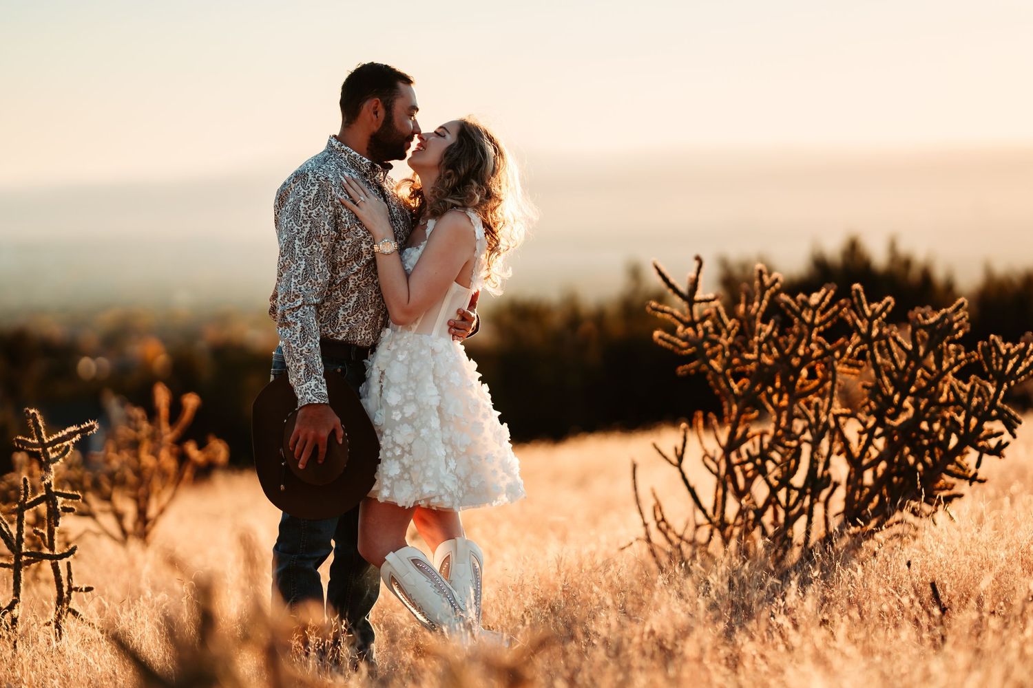 Romantic couple embraces in golden field at sunset while surrounded by dried plants and warm light.