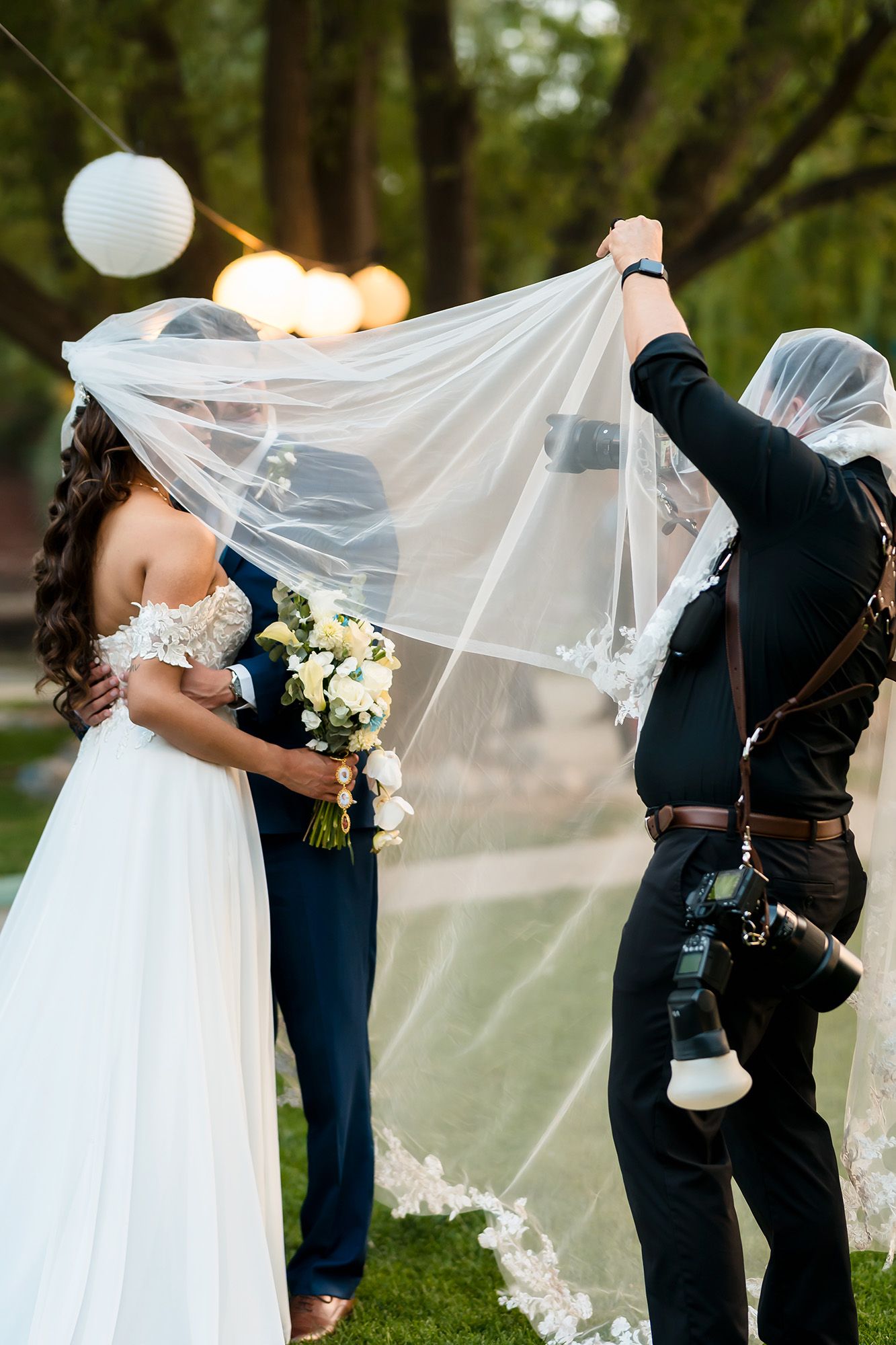 Wedding photographer lifting bride's veil while couple embraces outdoors, with lanterns and trees in background.