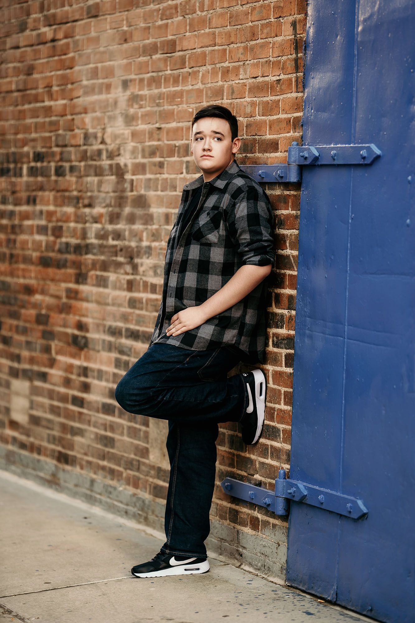 Young man in plaid shirt leaning against brick wall near blue door, wearing dark jeans and Nike sneakers.