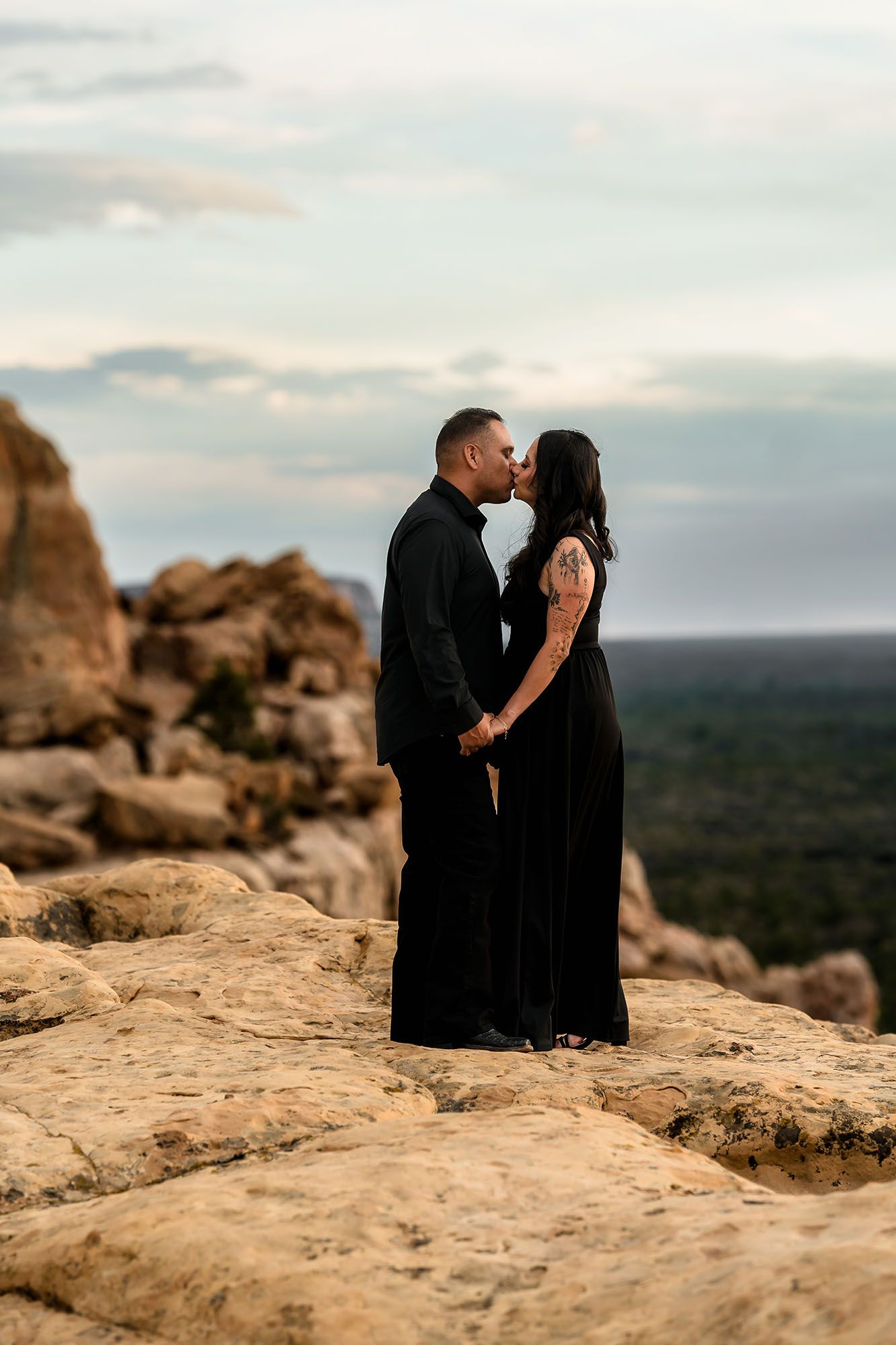 Couple in black attire kissing on rocky desert overlook at sunset with vast landscape behind them.