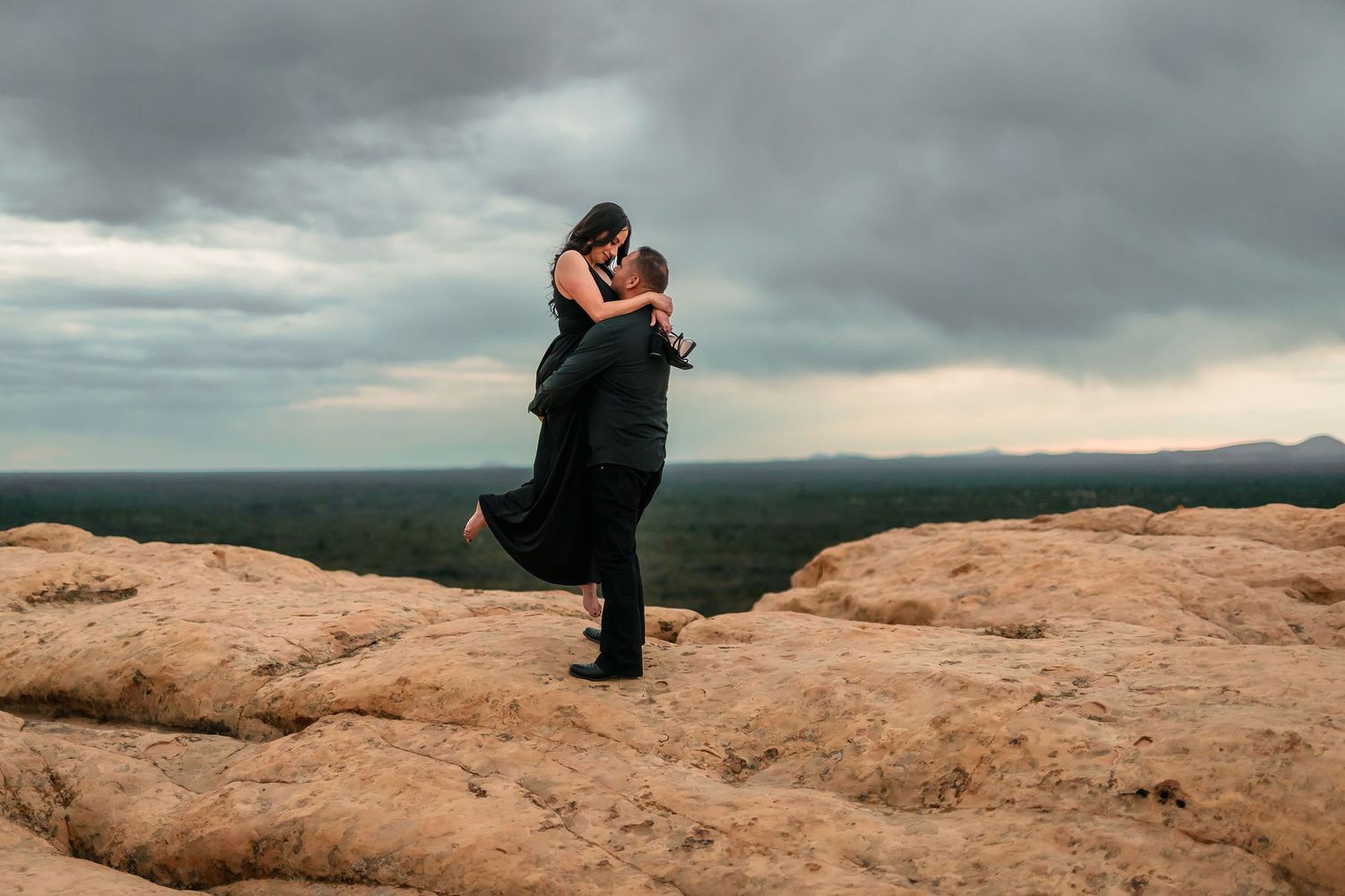 Couple embracing as man lifts woman on rocky desert overlook under moody storm clouds.