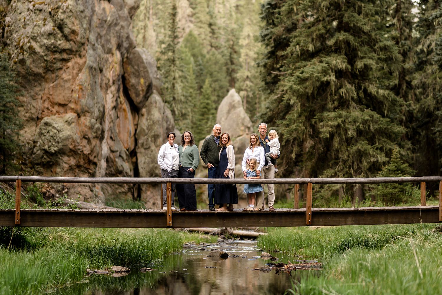 Family group portrait on wooden bridge surrounded by pine trees and rocky cliffs in a scenic mountain forest.