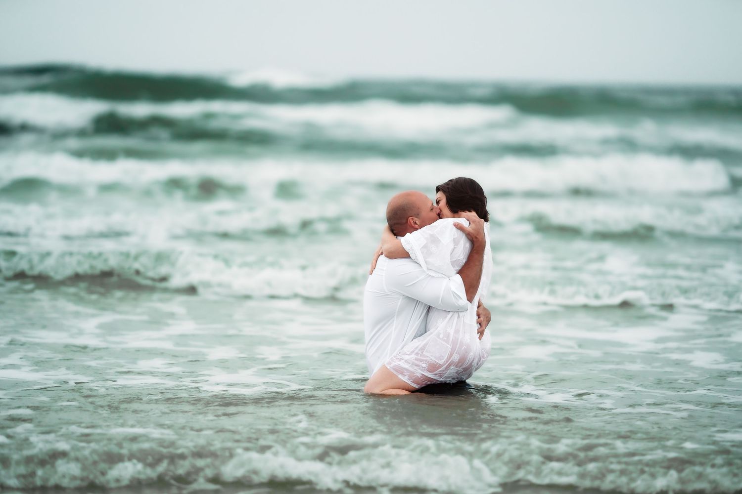 A couple in white clothing poses dramatically by ocean waves on a beach.