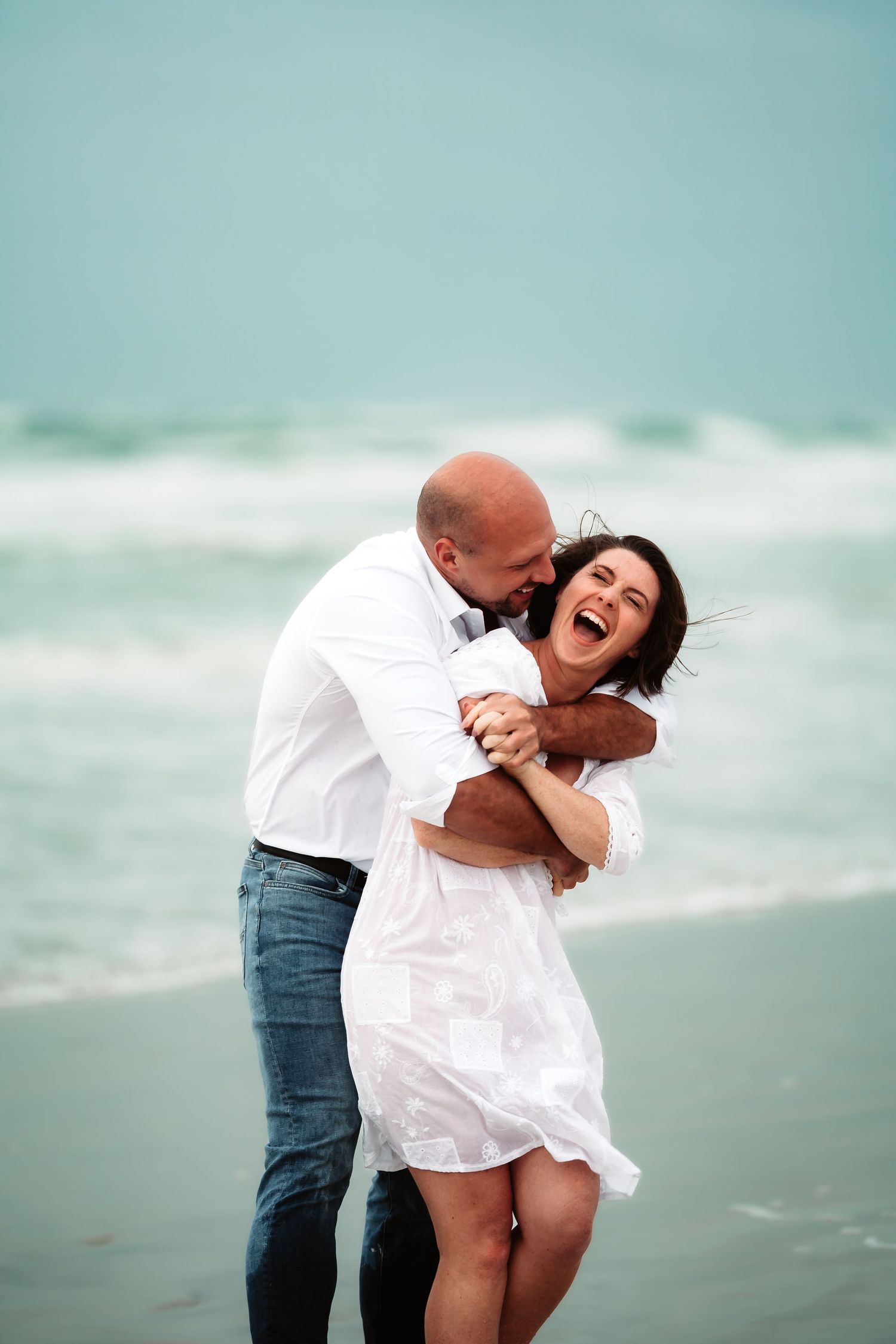 A couple shares a playful moment on the beach, laughing and embracing against a serene turquoise ocean backdrop.