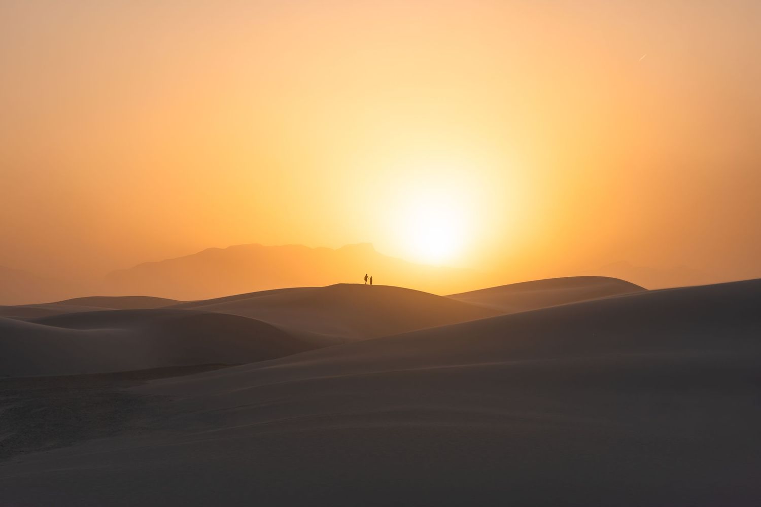 Silhouette of a person standing on desert sand dunes during a golden sunset over rolling hills.