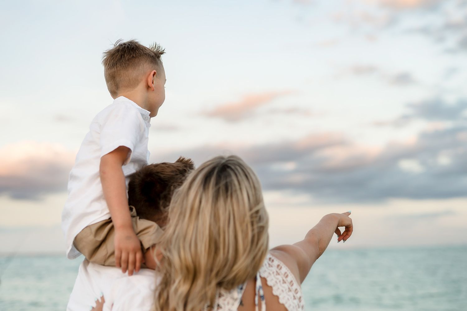 Mother holds toddler son on her shoulders at beach sunset, pointing at ocean view.