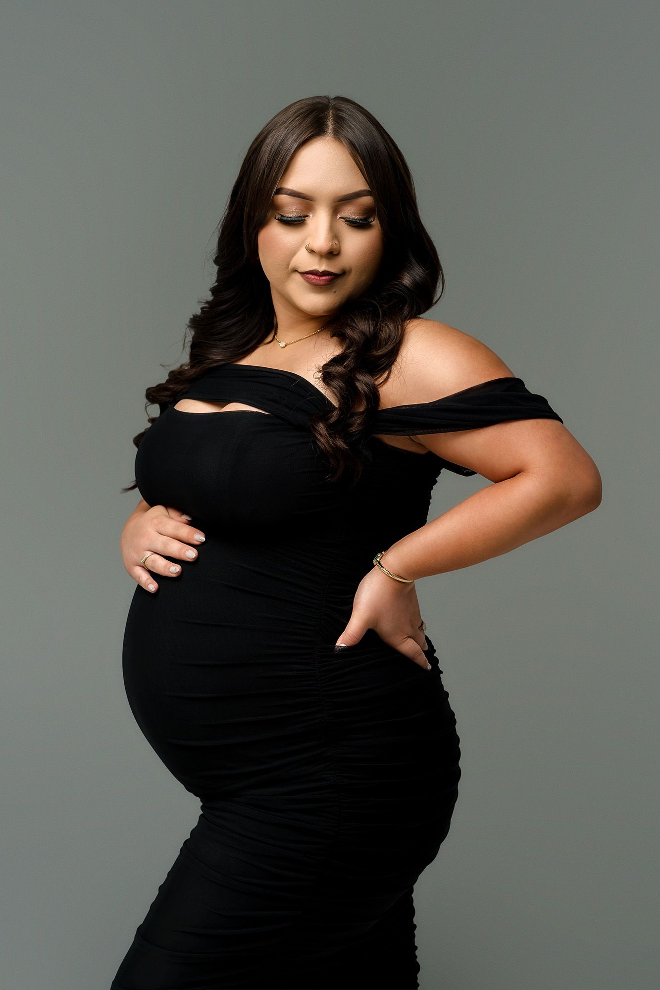 Pregnant woman in black off-shoulder dress posing gracefully, cradling baby bump in professional maternity photo shoot.