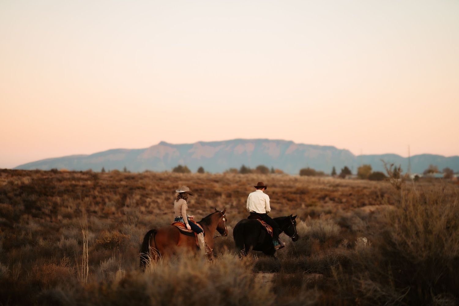 Two riders on horseback traverse a rugged desert landscape at sunset with mountains in the distance.