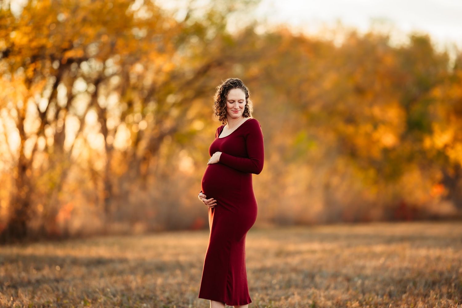 A person in a burgundy velvet dress poses in a golden autumn field during sunset.