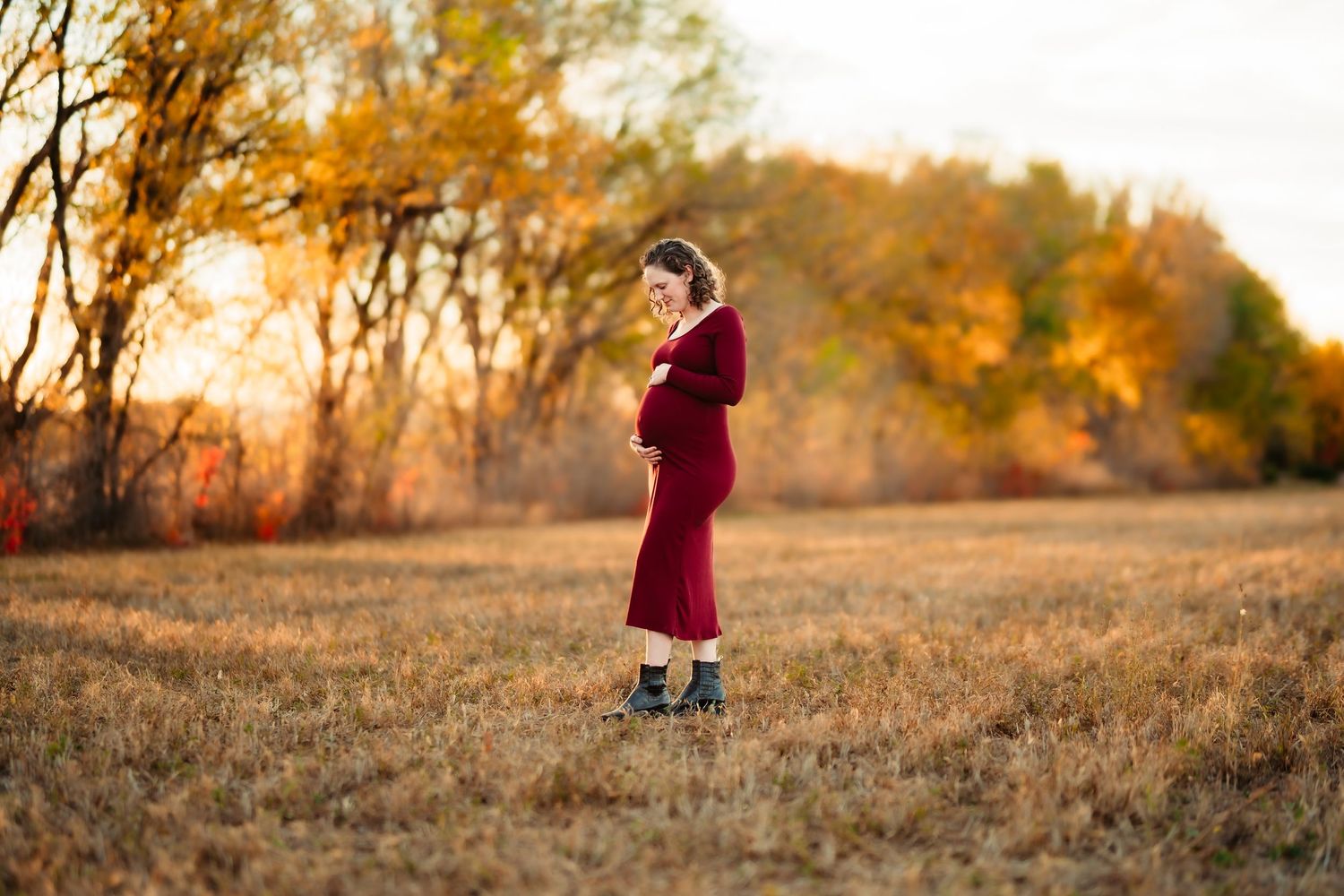 A child in a burgundy dress walks through a golden autumn field at sunset with warm glowing light.