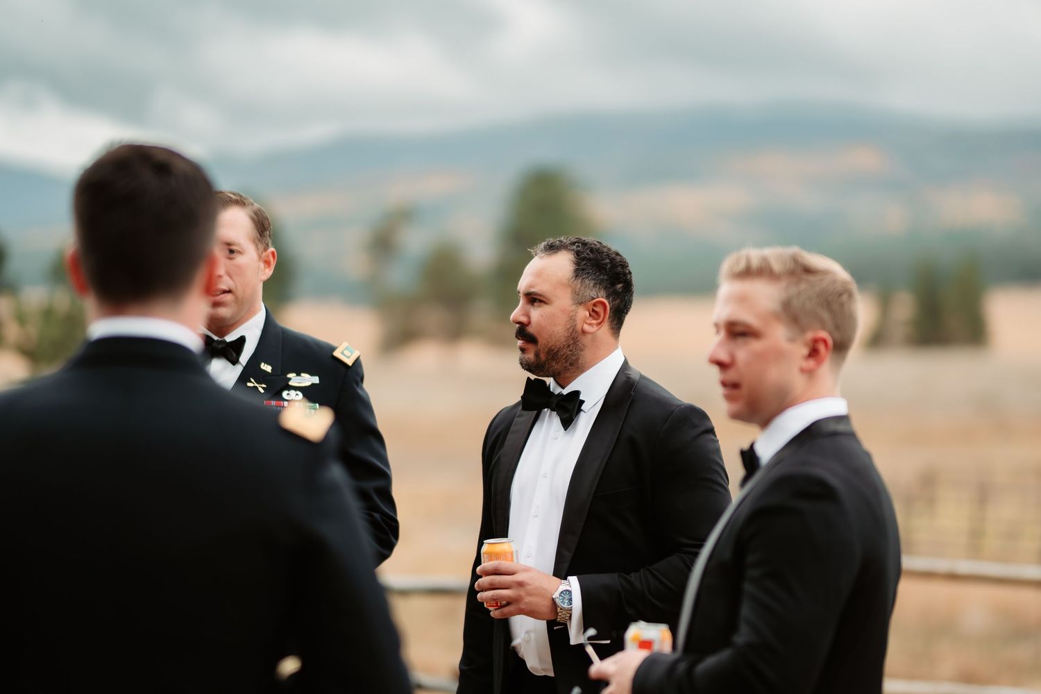 Groomsmen in black tuxedos celebrate and toast with drinks at an outdoor wedding reception.