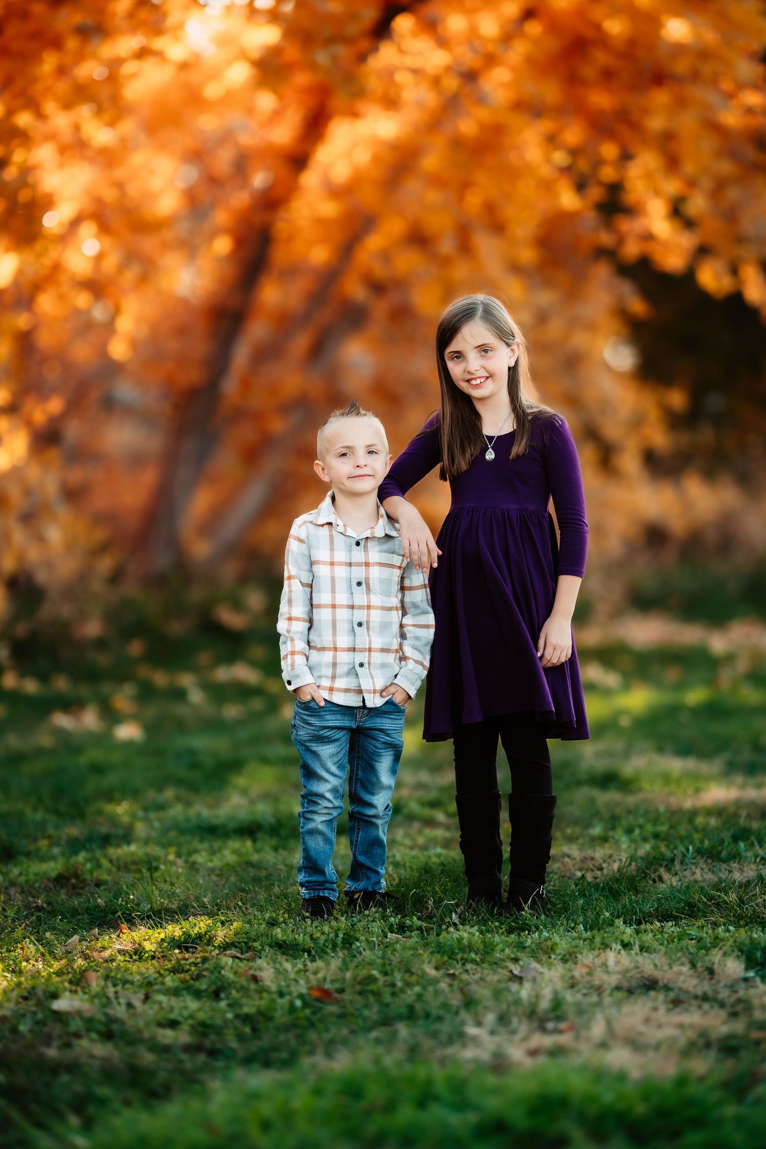 Two children stand together on grass against vibrant orange autumn foliage in the background.