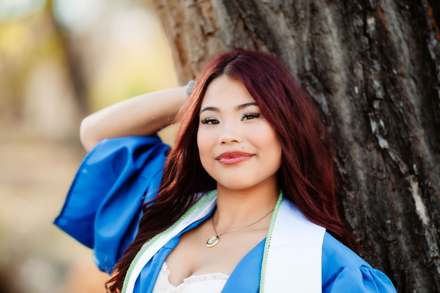 A woman in a bright blue graduation gown stands against a tree trunk with autumn foliage in the background.
