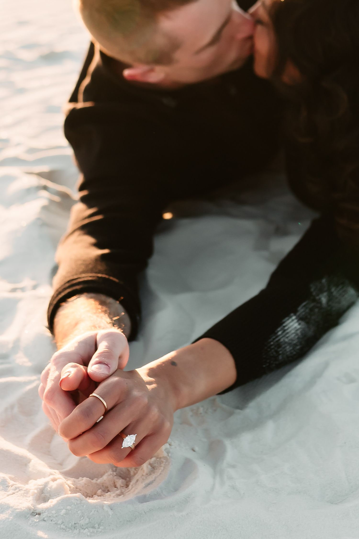 Close-up of clasped hands with wedding rings resting on white sand in warm sunlight.