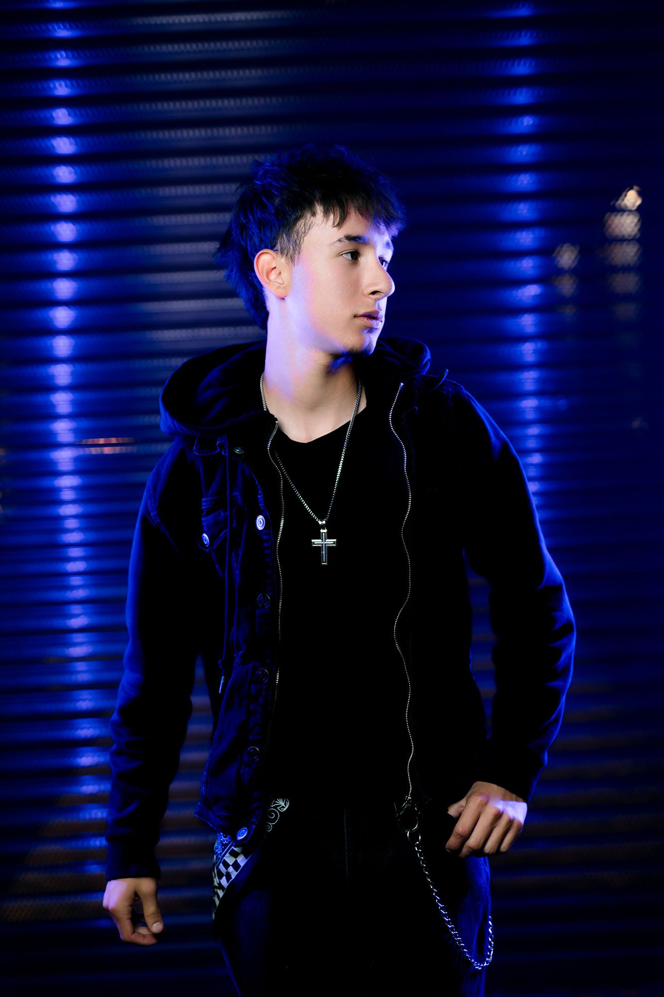 Young man in black hoodie with cross necklace posing against blue-lit metal wall at night.