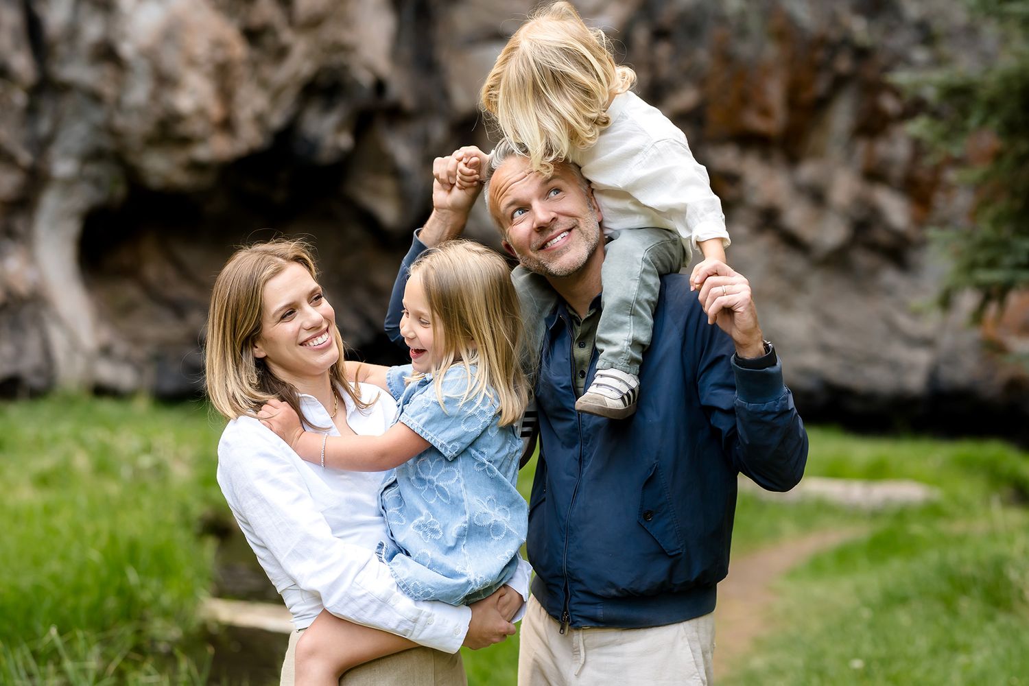 Happy family of four posing outdoors near rocky cliffs, parents holding young children while smiling in a lush green setting.