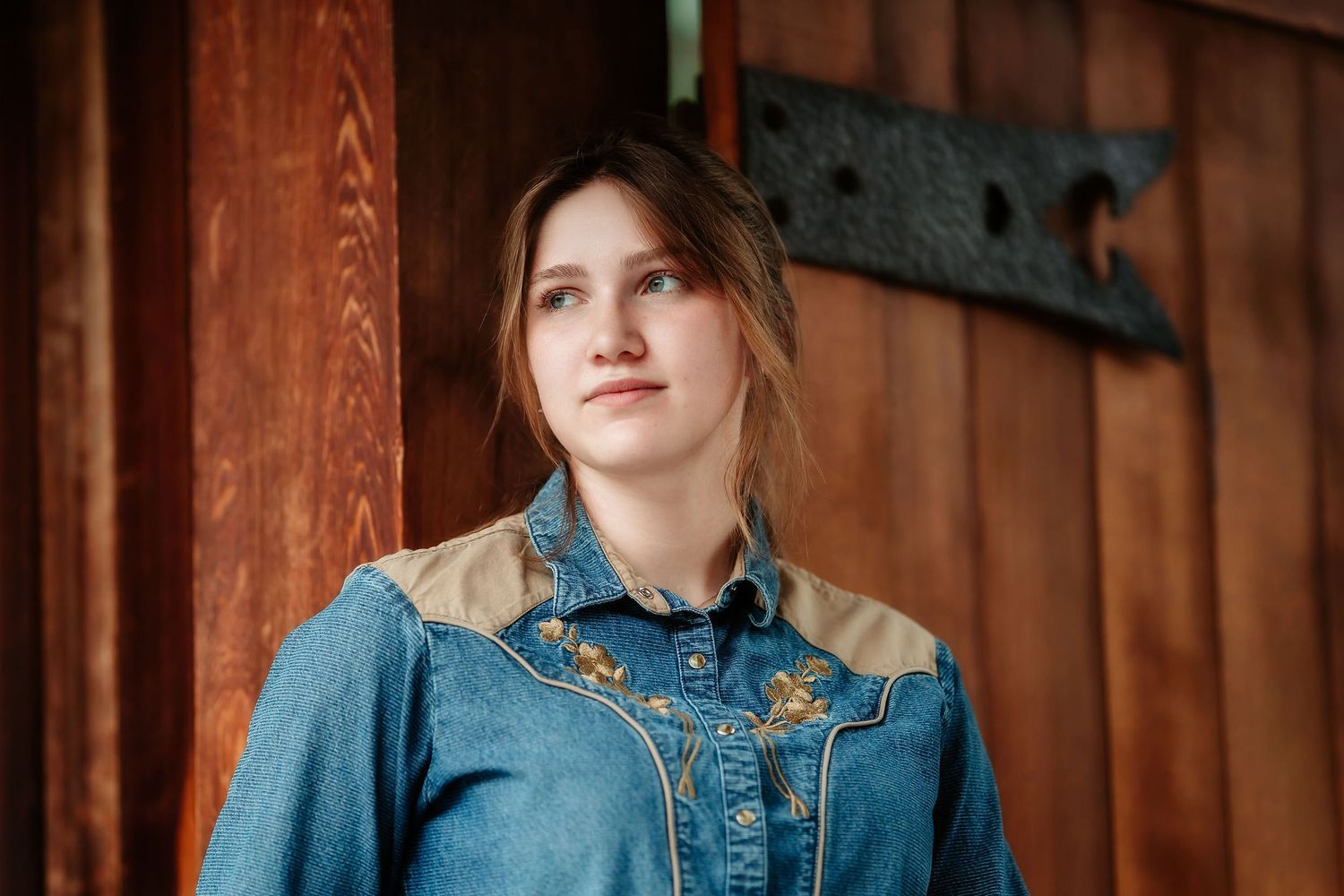 Young woman in embroidered denim western shirt leaning against rustic wooden barn wall looking upward.