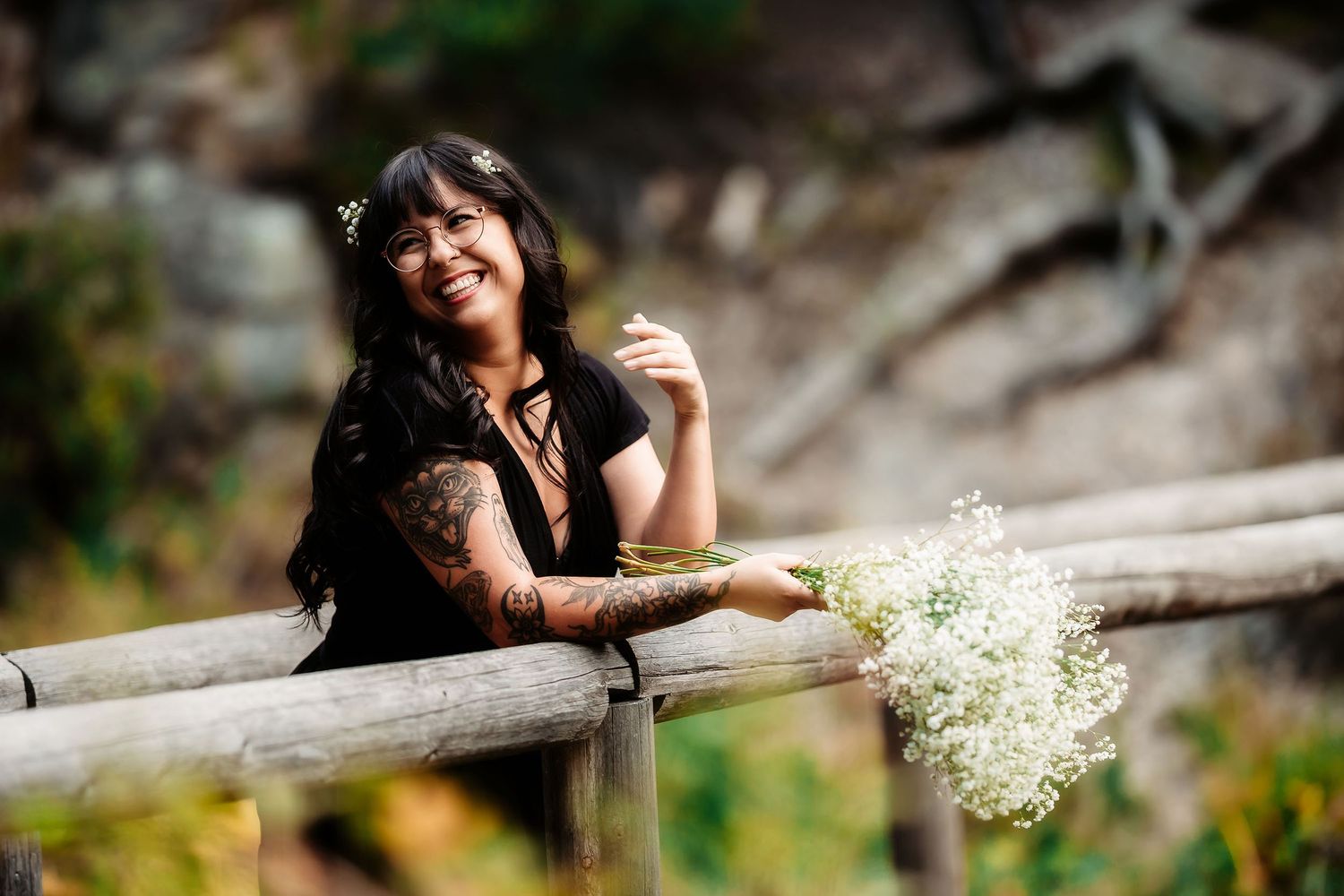 Smiling tattooed woman in black top holds white wildflowers while leaning on a wooden fence.