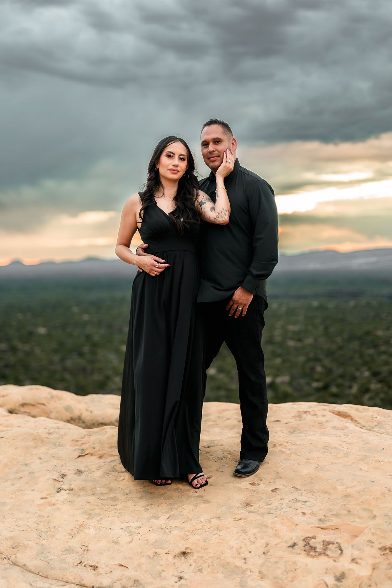 A couple poses on a sandstone cliff at sunset, dressed in matching black outfits with a stormy sky backdrop.
