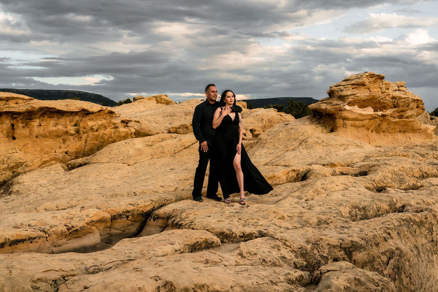 Couple in black formal attire posing on golden sandstone rock formations under dramatic cloudy skies.