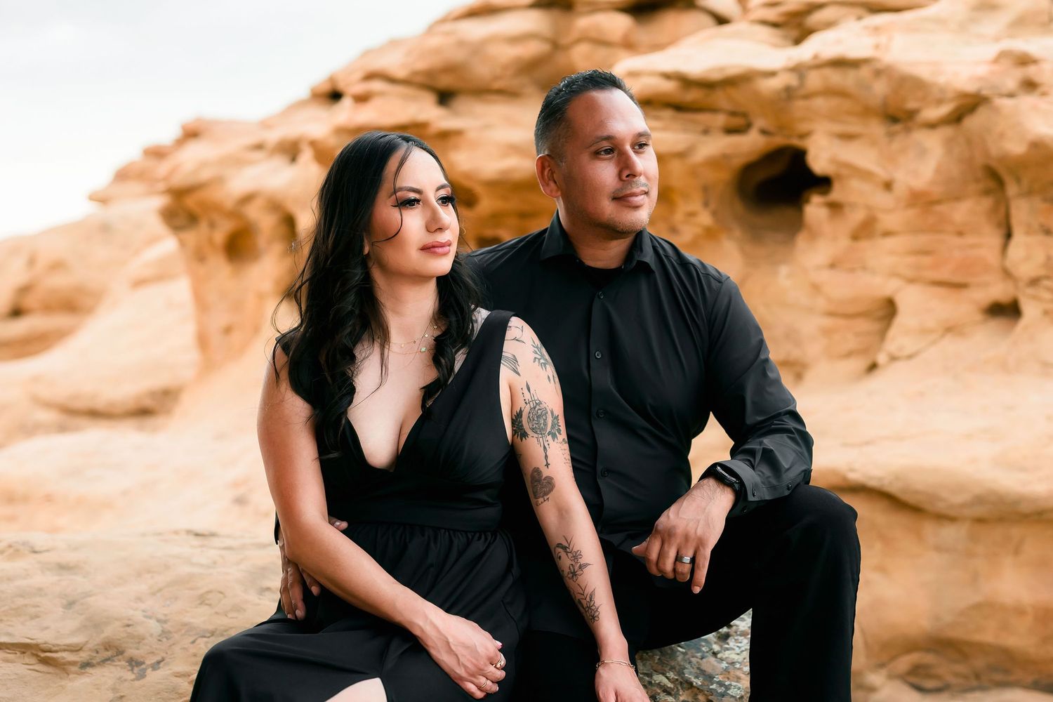 Couple in matching black outfits sitting on desert rocks with sandstone formations backdrop.