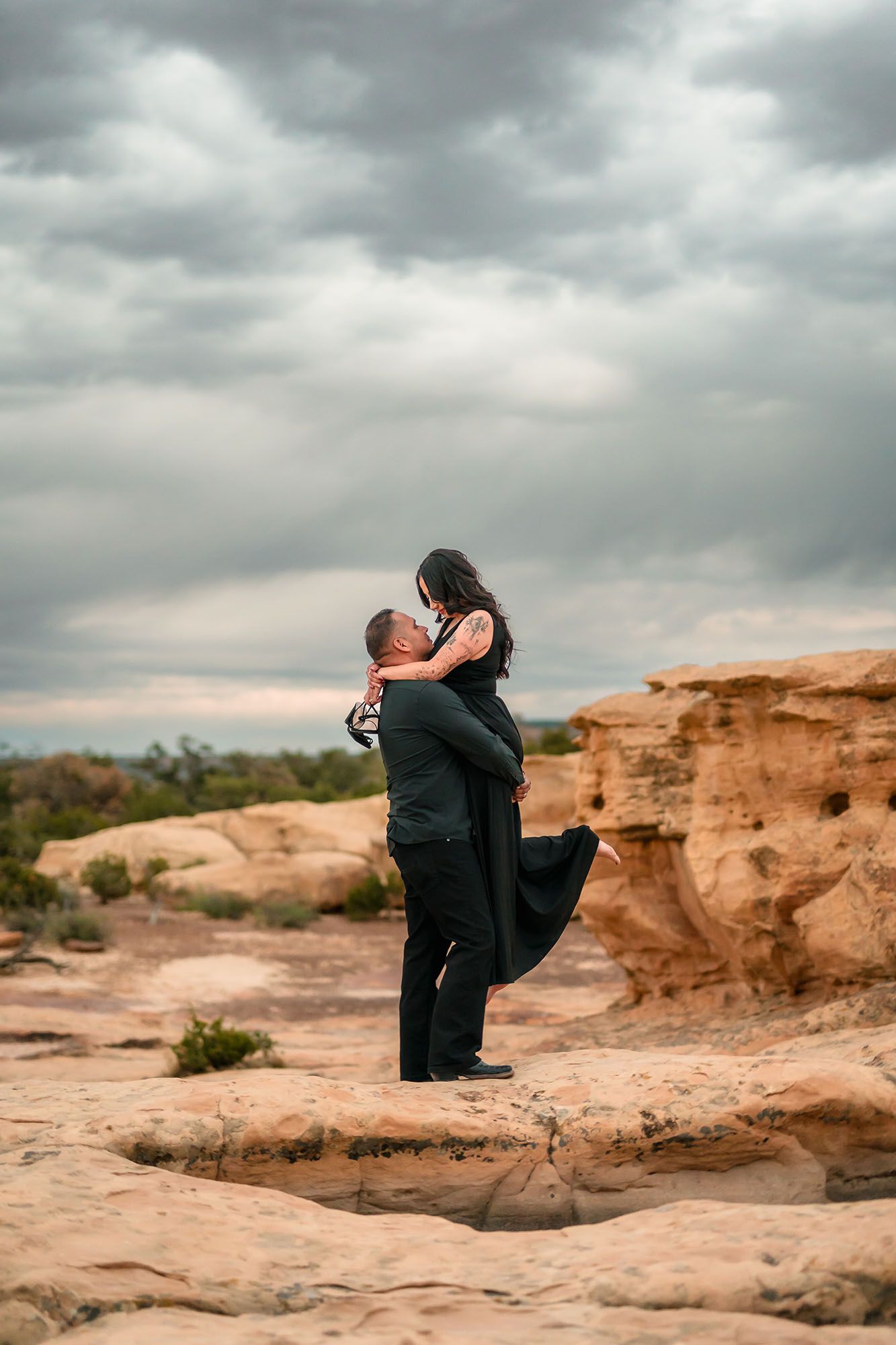 Couple embracing on red rock formation under dramatic cloudy sky in desert landscape.
