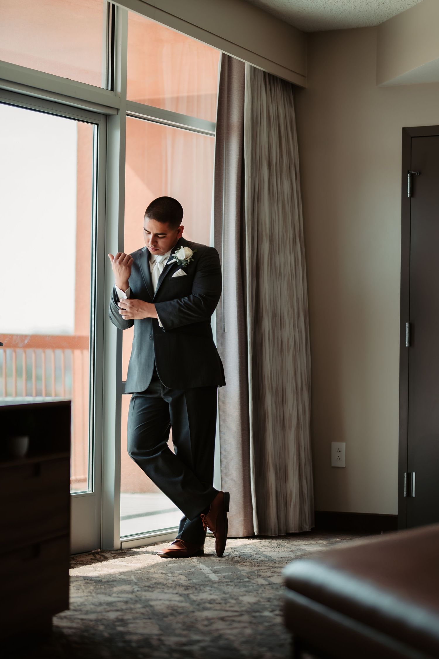 A figure in a dark suit poses expressively against large windows in an elegant room with dramatic lighting.