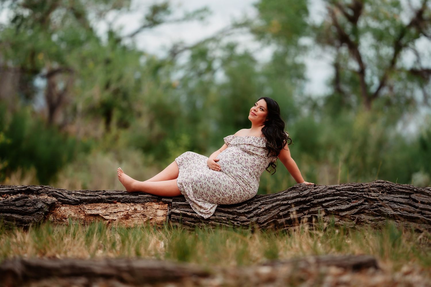 A woman in a white floral dress lying gracefully on a rustic wooden log in an outdoor nature setting.