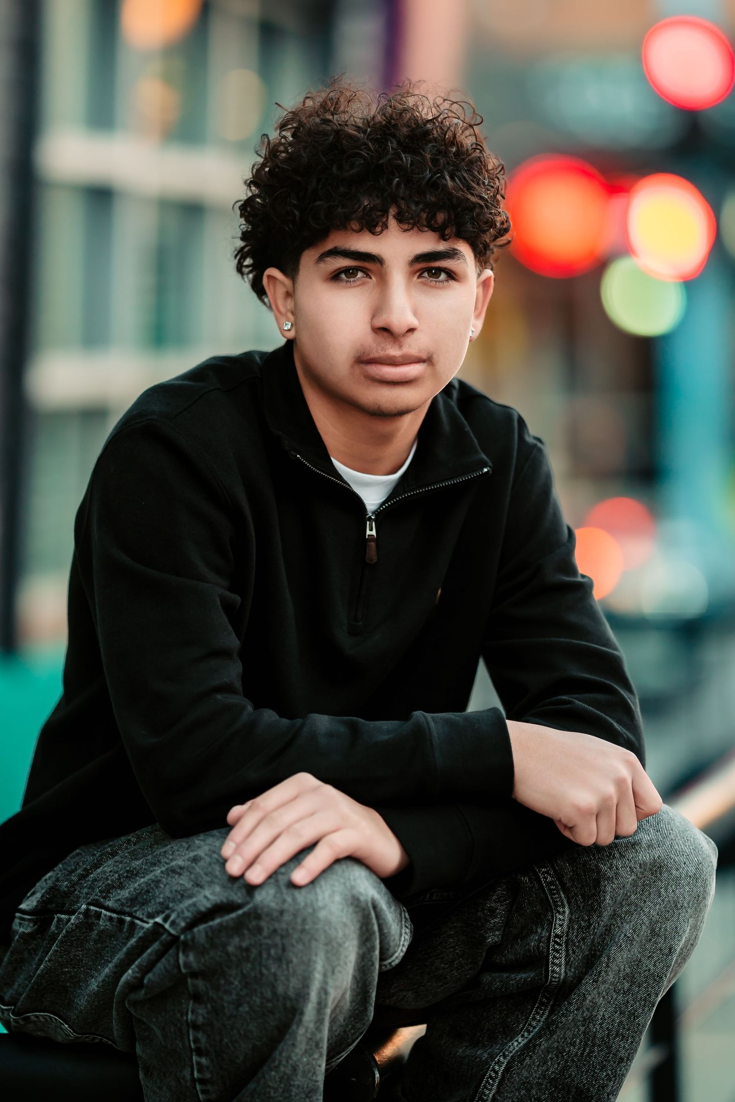 Young person in black hoodie sits casually on urban street with blurred city lights in background.